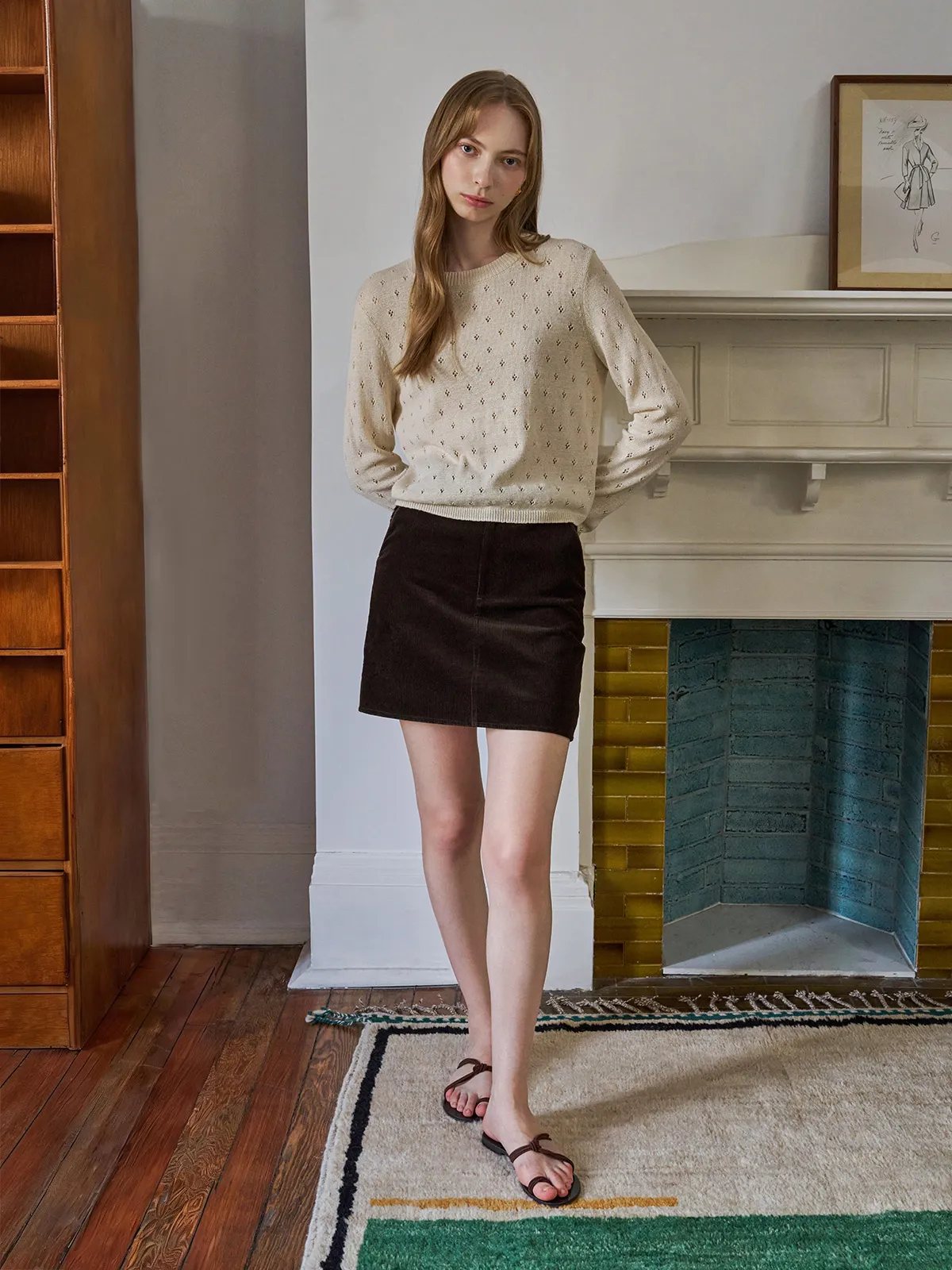 Model in cream knit sweater and brown DENVER skirt standing beside fireplace with patterned rug