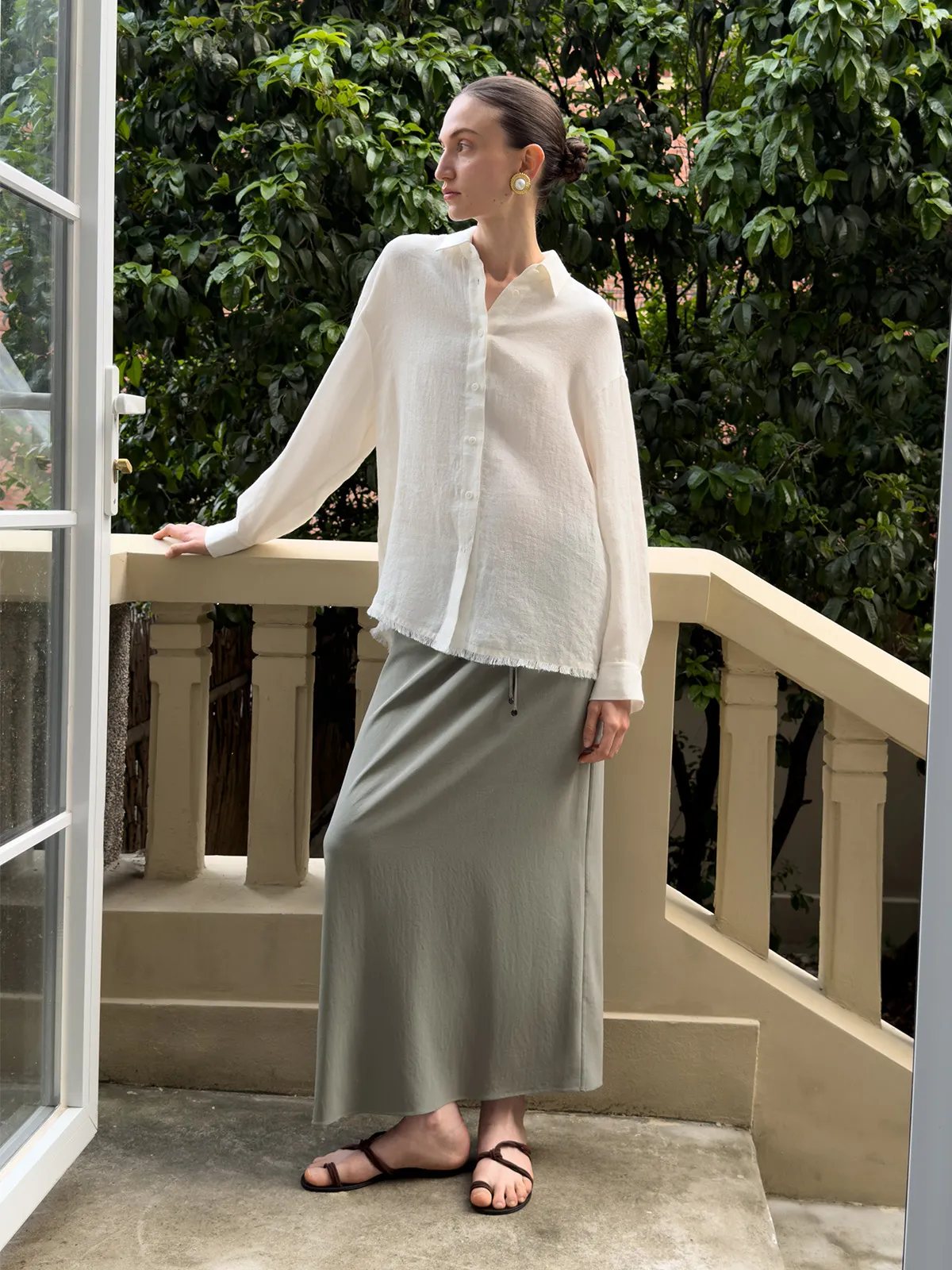 Model on balcony wearing KATRINA midi skirt with oversized white linen shirt and gold earrings