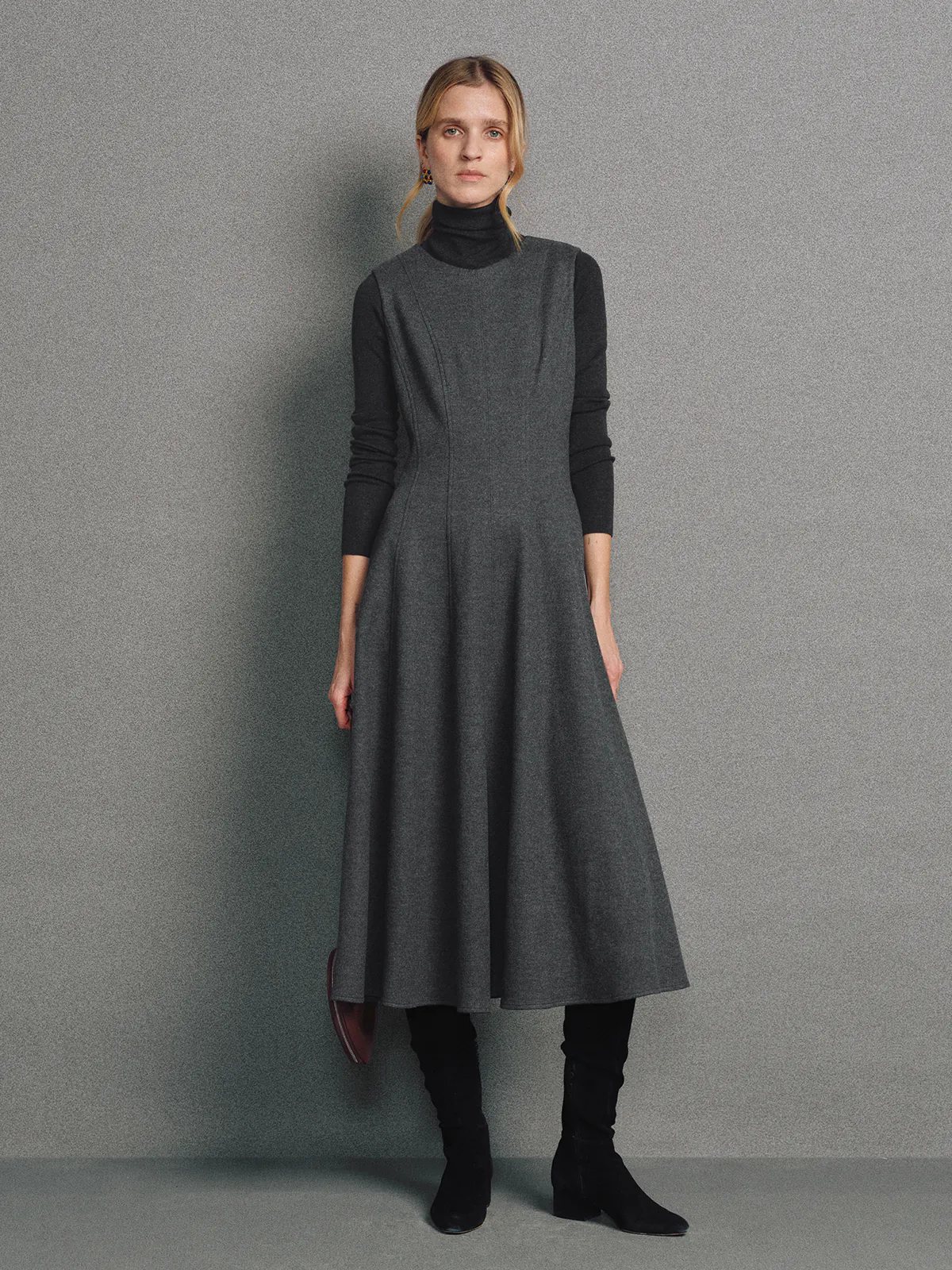 Model wearing a sleeveless dark gray wool knit A-line dress over a turtleneck with boots.