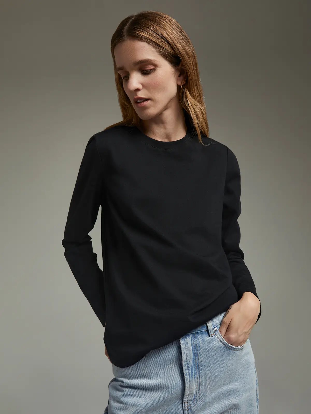 Model wearing a black long-sleeve crewneck T-shirt with one hand in pocket of light-wash jeans, looking downward.