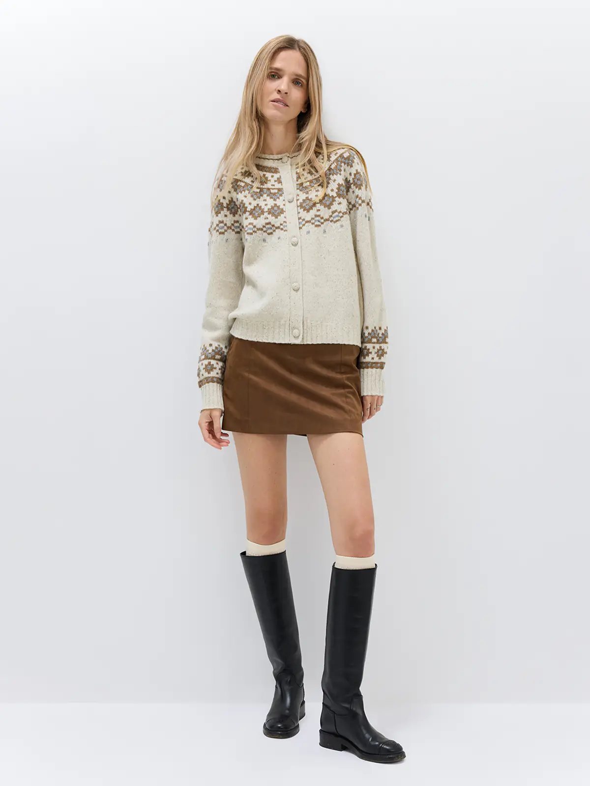 Model wearing an Oatmeal Fair Isle cardigan with geometric patterns, layered over a turtleneck and trousers, holding a bag.