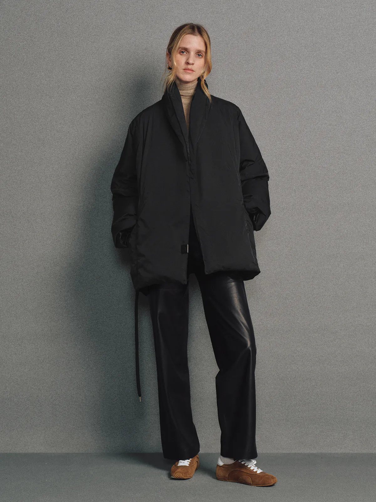Side view of the Black OPHELIA relaxed wrap-style down coat with voluminous sleeves and tied waist, hands in pockets.