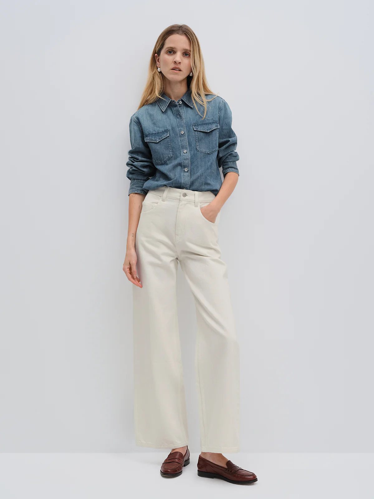 Front view of model wearing white wide-leg jeans with denim shirt and loafers, hand in pocket.