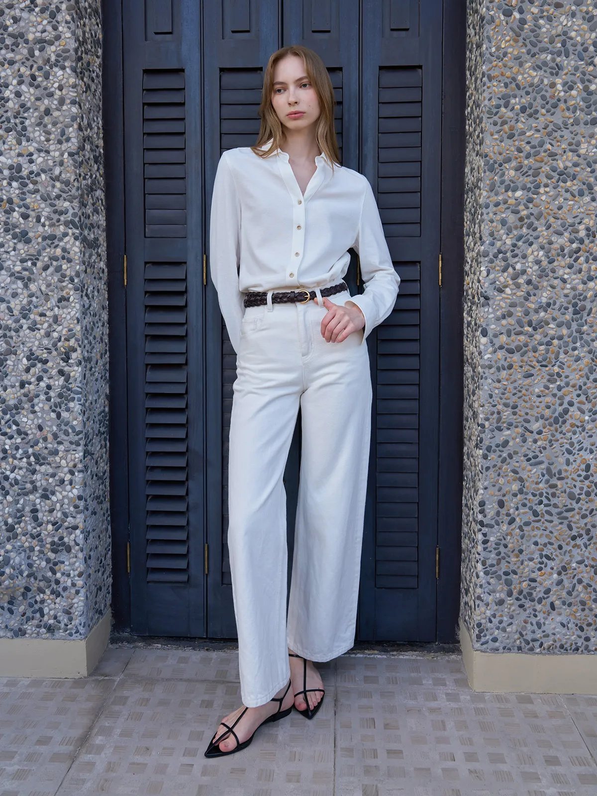 Model in white shirt and cream wide-leg pants wearing PLAIT belt against pebble wall and black shutters
