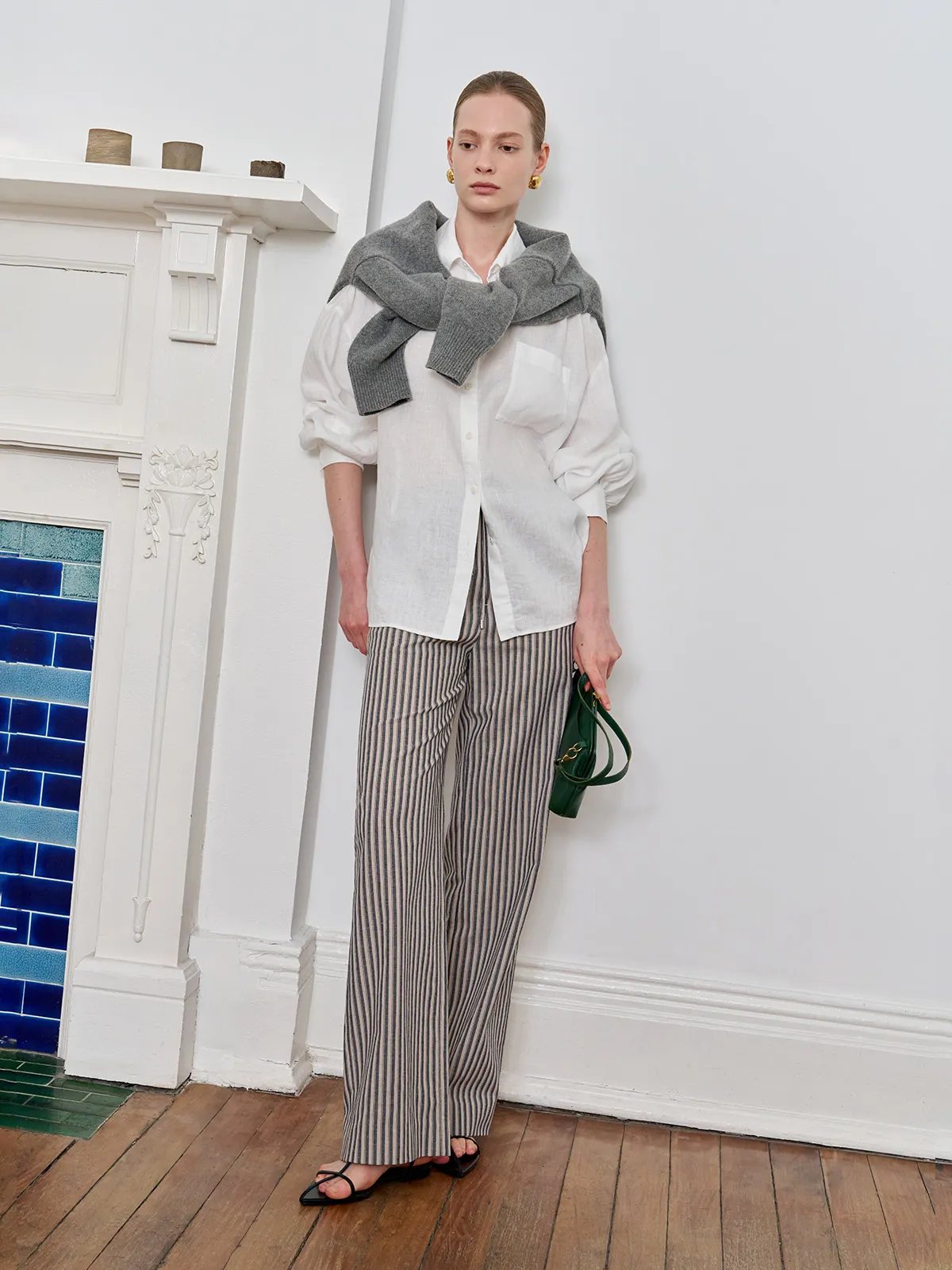 Model styled with white linen shirt, grey sweater draped shoulders, green mini bag, and WHIMSY striped trousers