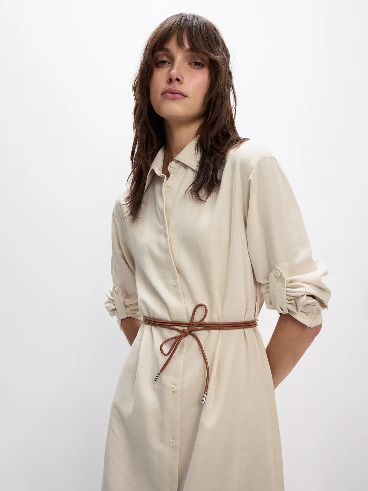 Side view of the Natural JOANNA shirt dress with rolled sleeves, button-front placket, self-fabric tie belt, and hands in pockets.