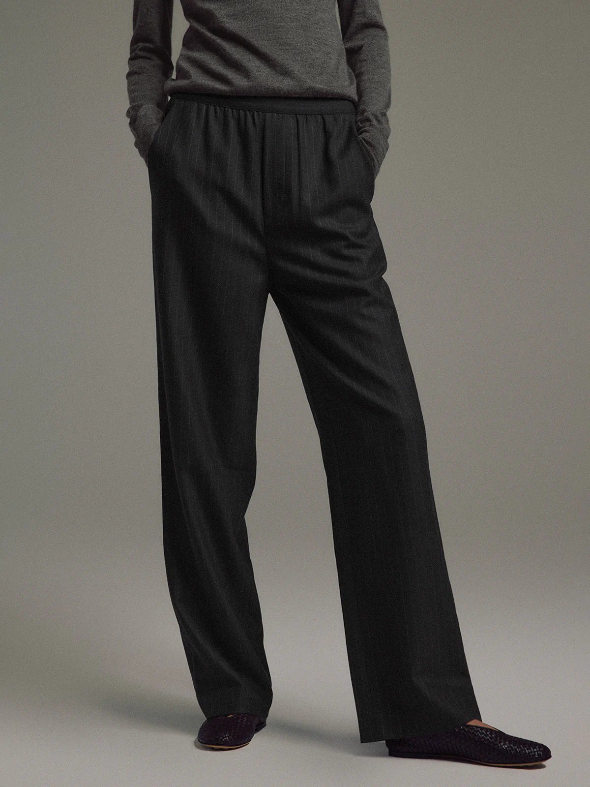 Side view of model in Black/White wool-blend straight trousers with elastic waist, hands in pockets, wearing a top and dark woven slippers.