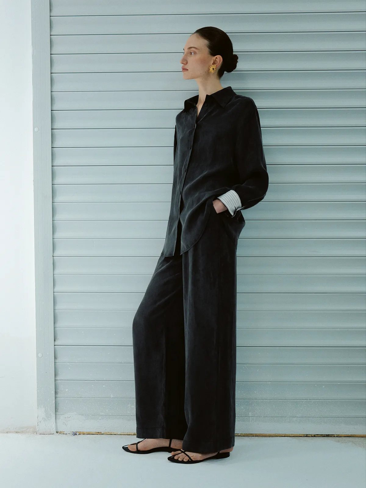 Side profile of model in black NOBU Trousers and matching shirt against grey roller shutter wall