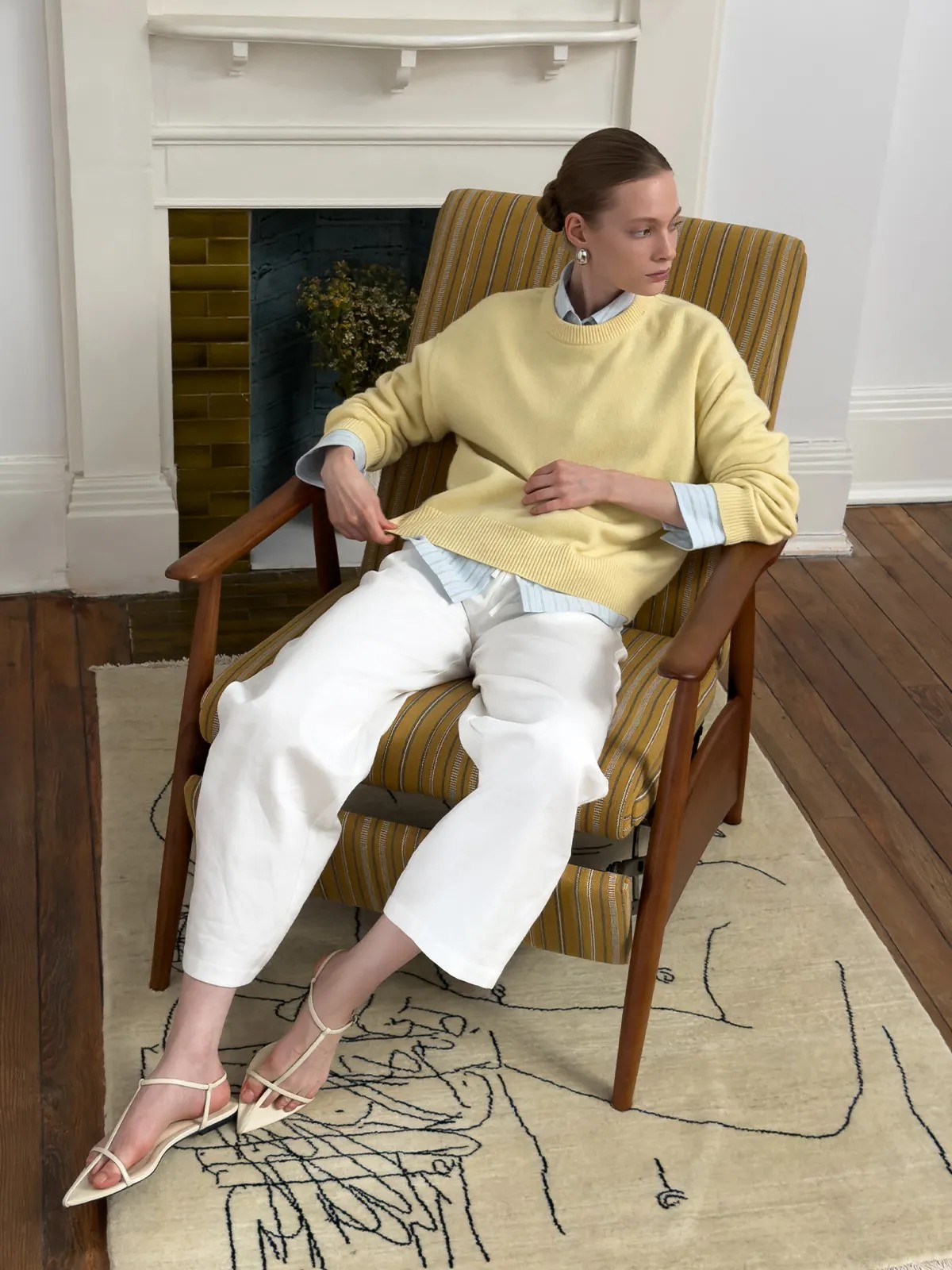 Model seated in mid-century chair wearing white LEIGHTON trousers with yellow sweater and striped shirt, showing cropped hem and drape