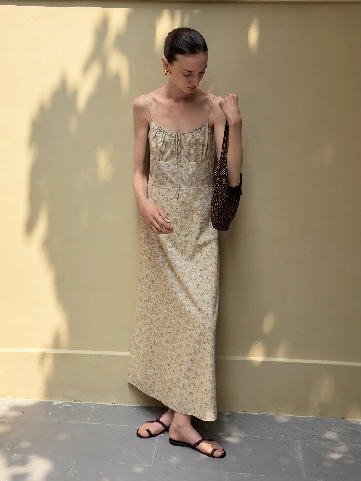Model in beige floral PIPPA dress looking down, holding beaded bag against sun-shadowed wall, minimalist aesthetic