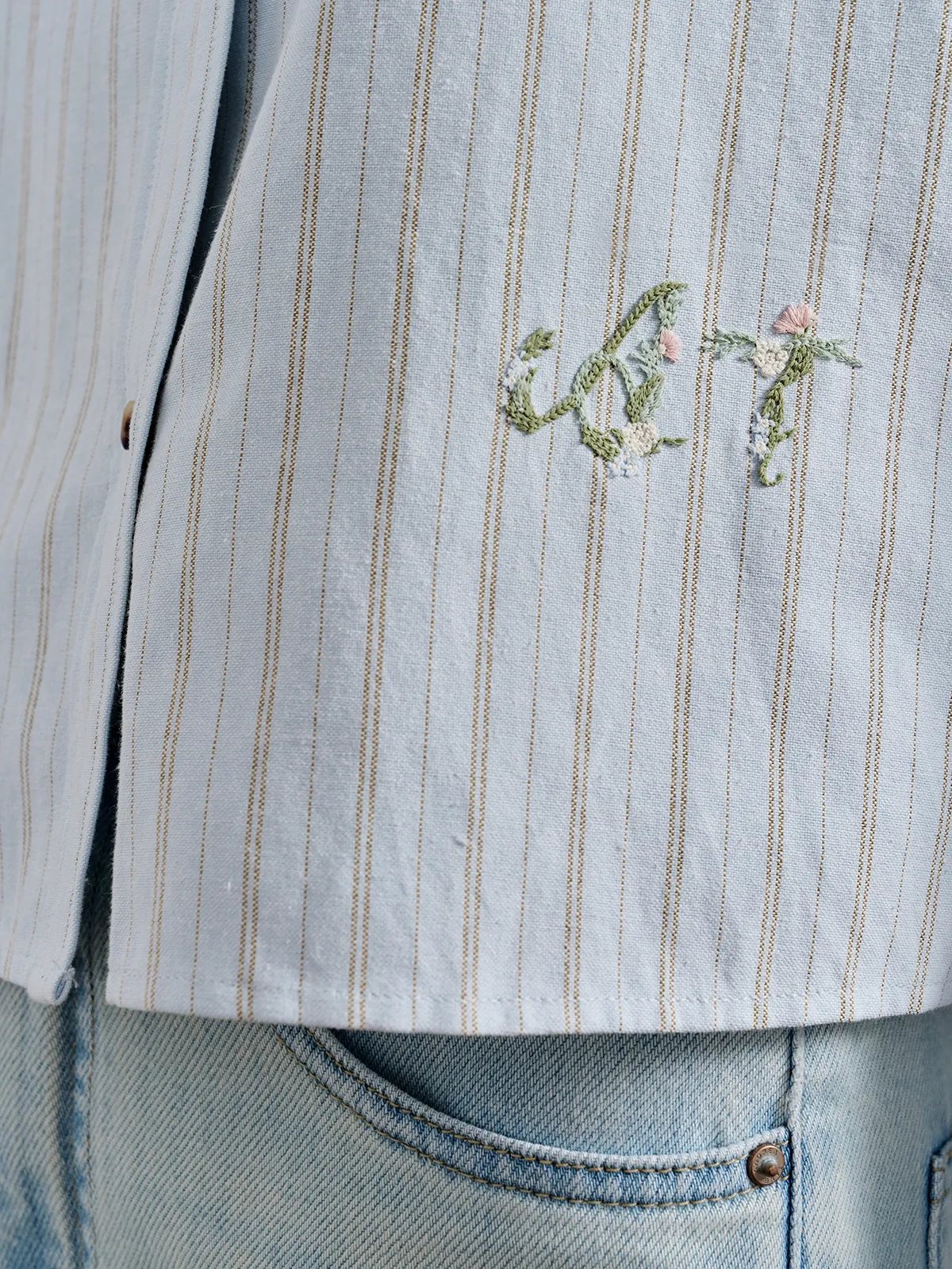 Close-up of botanical embroidery on the light blue striped TILLY shirt, with green stems, ivory flowers and pink buds