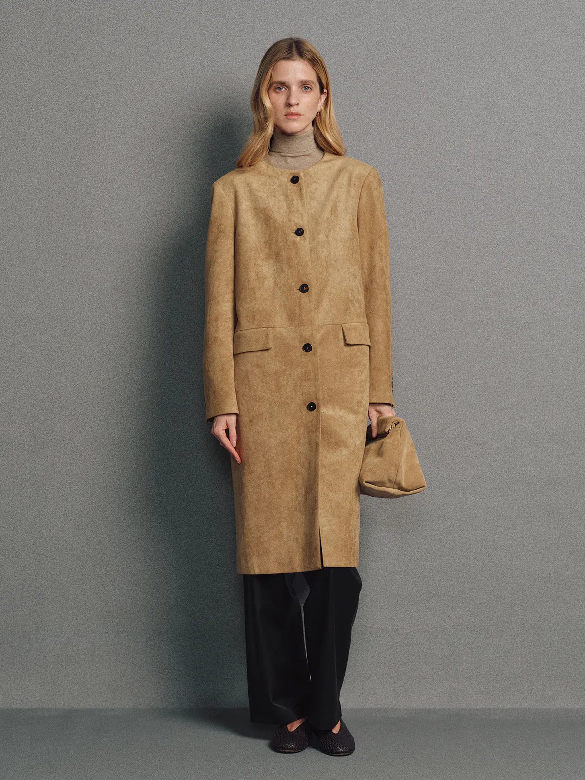 Front view of model wearing a camel faux suede collarless long coat with front buttons, dark trousers, sneakers, and a bag.