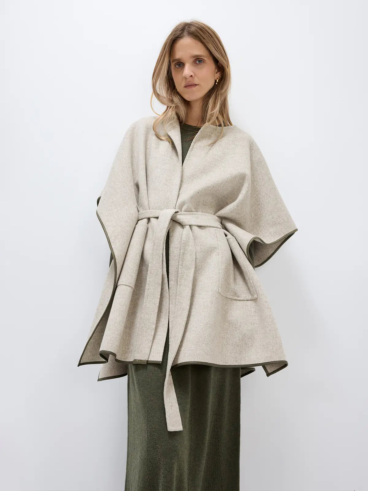 Model in Beige cape coat with belt tied, hands in pockets, holding a bag, paired with pants and loafers.