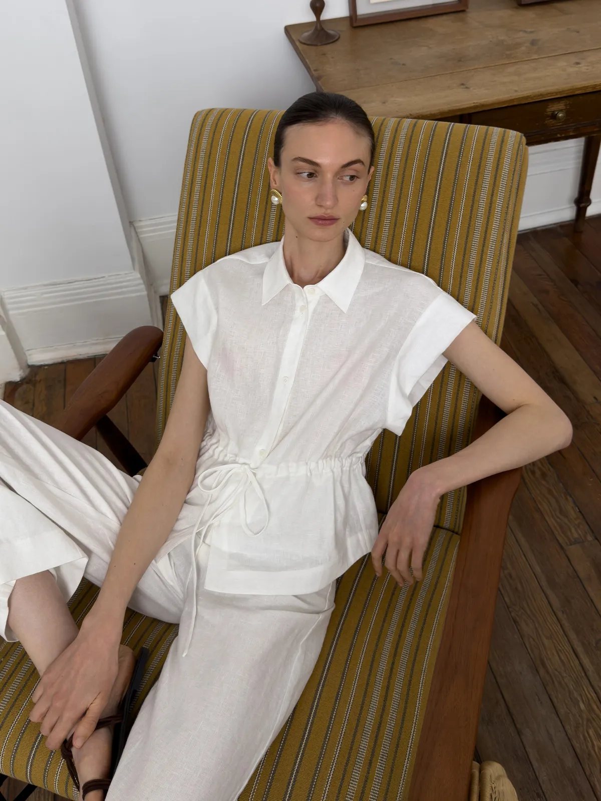 Model seated in striped armchair wearing MAREN white linen shirt with drawstring waist, paired with wide-leg pants and pearl earrings