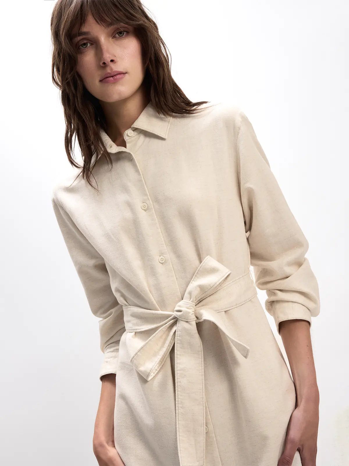 Side view of the Natural JOANNA shirt dress with rolled sleeves, front buttons, self-fabric tie belt, holding a woven bag.
