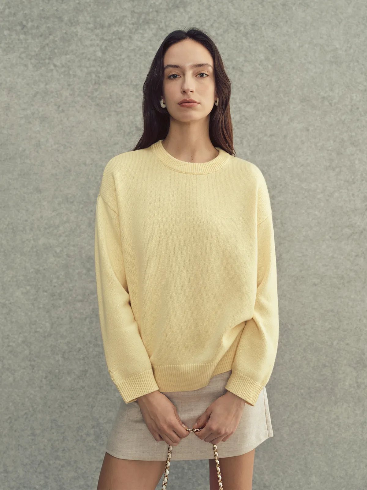 Front-facing model in pale yellow JILL jumper, hands clasped holding chain strap, neutral expression