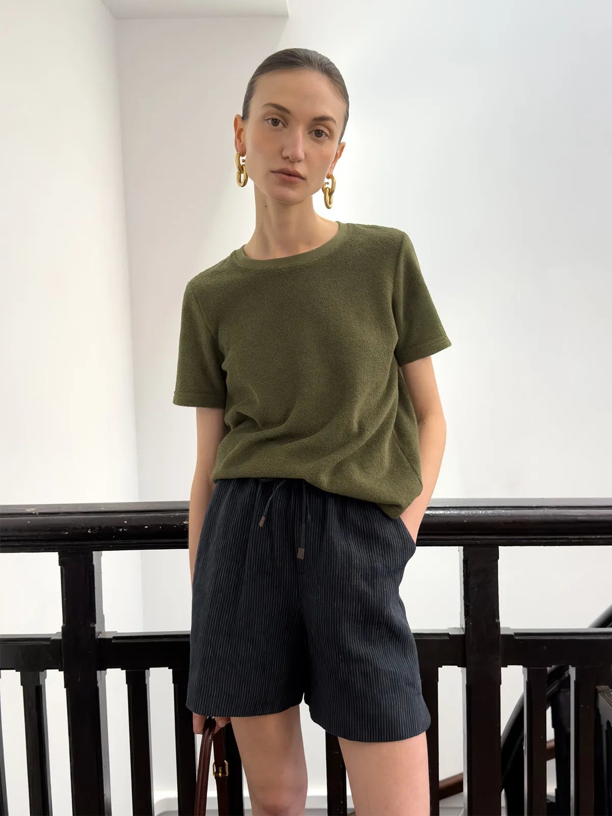 Front-facing model in olive tee and NEVAEH shorts, hands in pockets, against white wall and railing