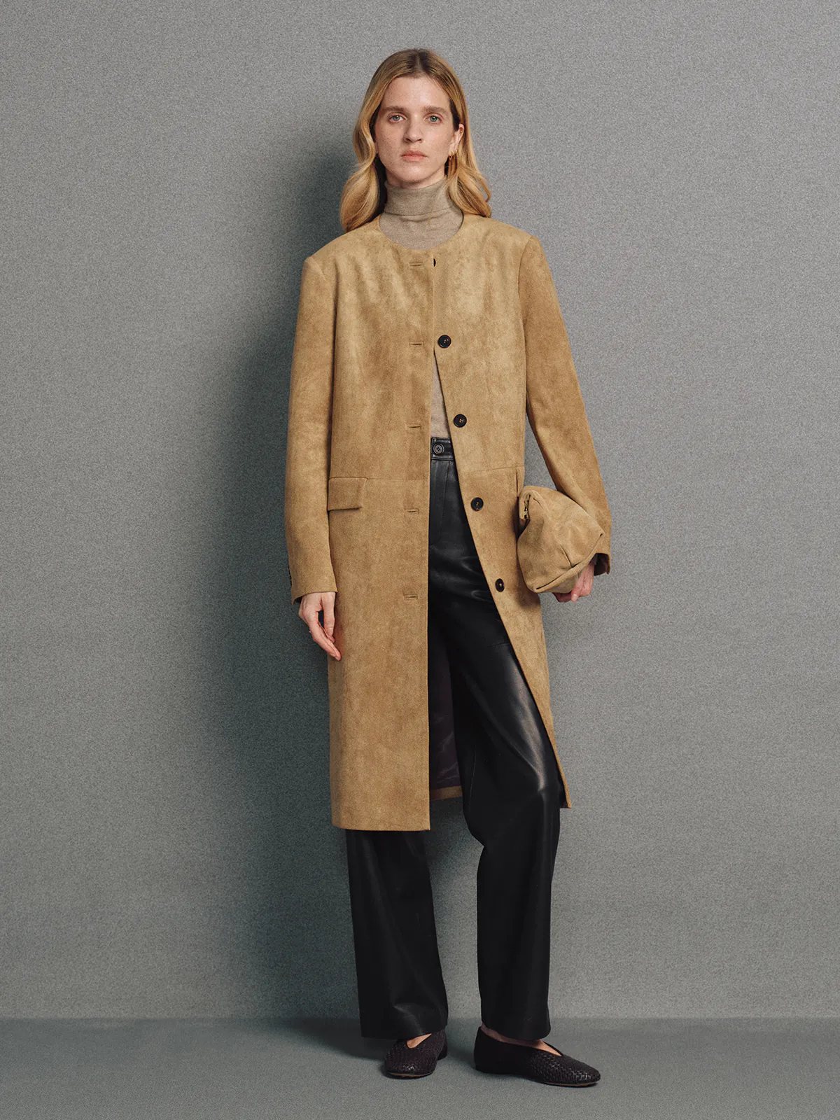 Front view of model wearing a camel faux suede collarless long coat with front buttons, paired with dark trousers and a turtleneck.