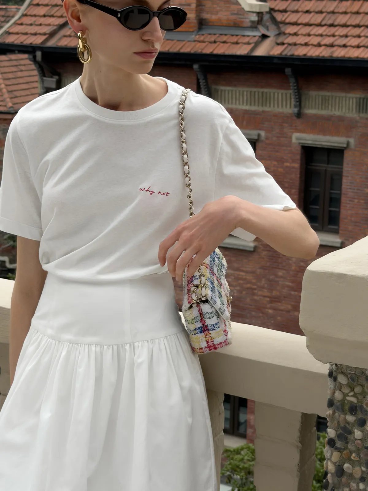 Model in white tee and white skirt holding colorful tweed mini bag, outdoor balcony setting