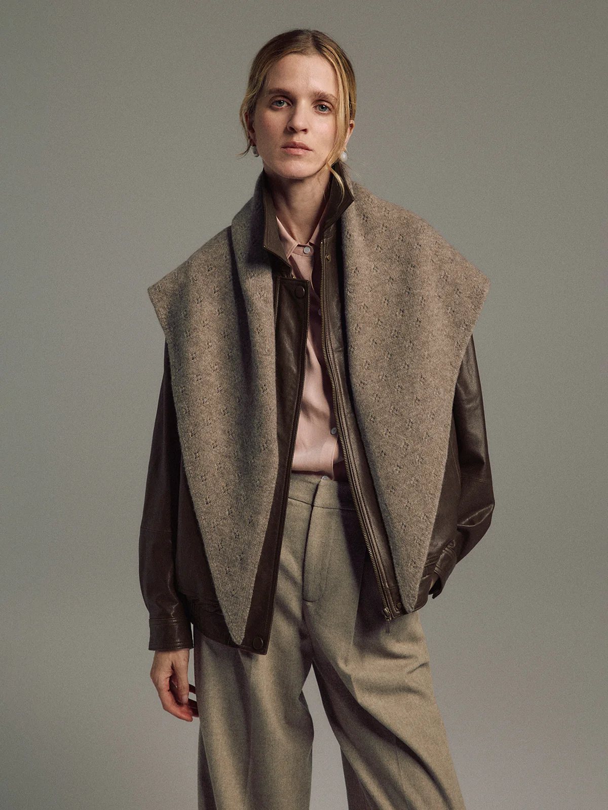 Model faces forward wearing the orange yak wool triangle scarf open over a cable-knit sweater and dark trousers.