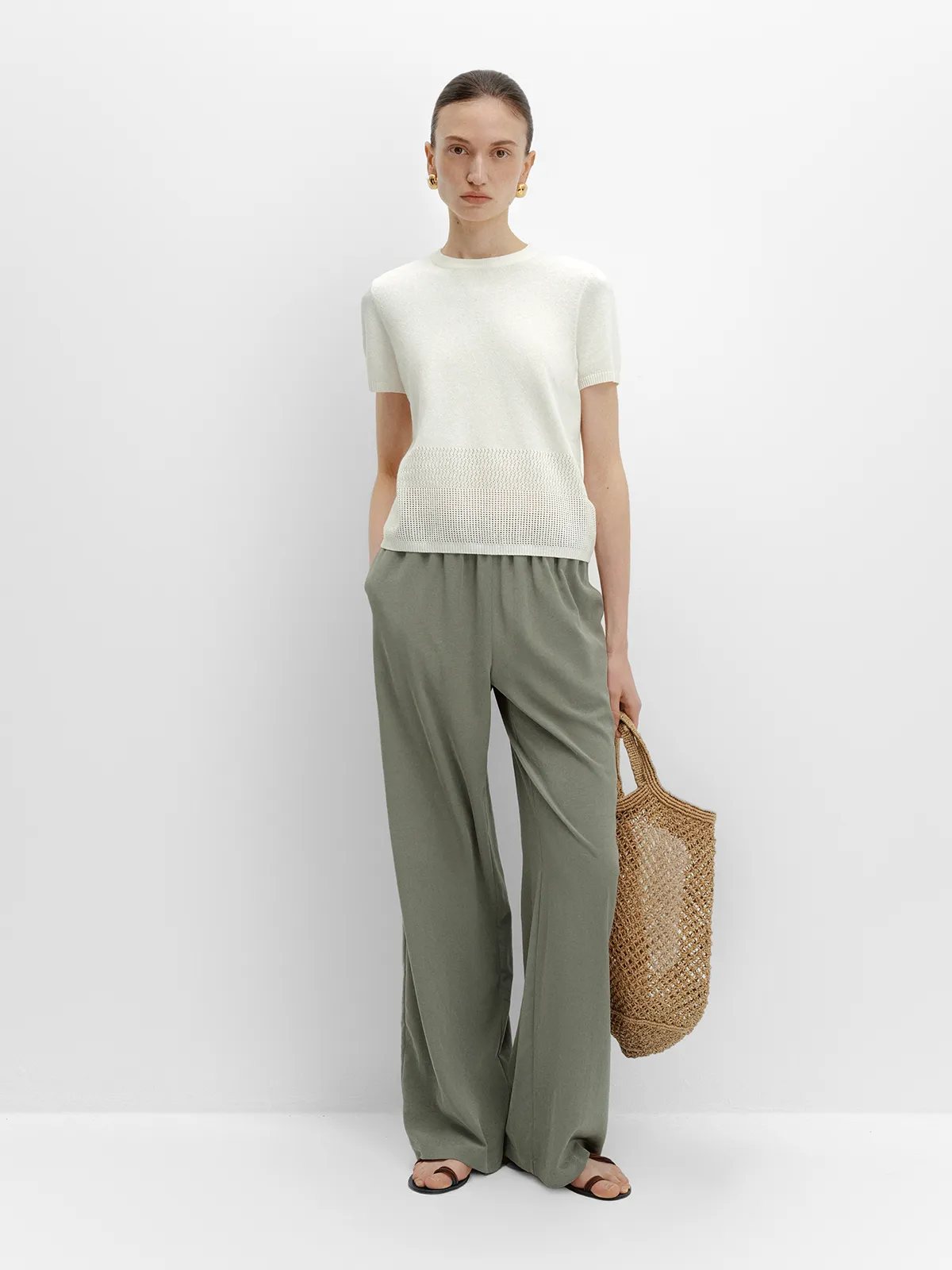 Model in andreatang PATRICIA trousers with cream knit top and woven straw tote bag
