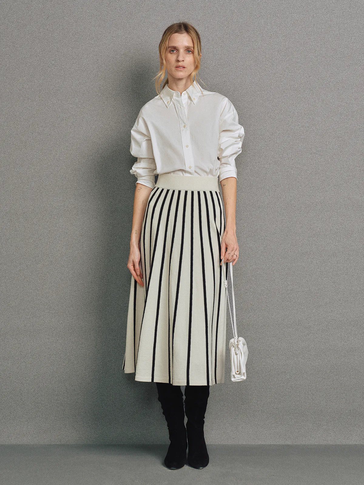 Front view of a model wearing a Black/White vertical stripe A-line knit skirt with white waistband, white shirt, and black boots.