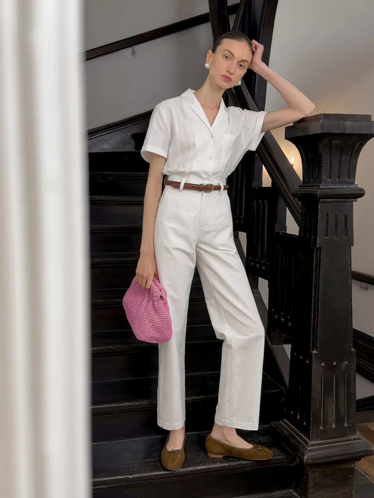 Model wearing white REID linen-cotton cropped trousers with short-sleeve shirt, brown belt, and pink crocheted bag on black staircase