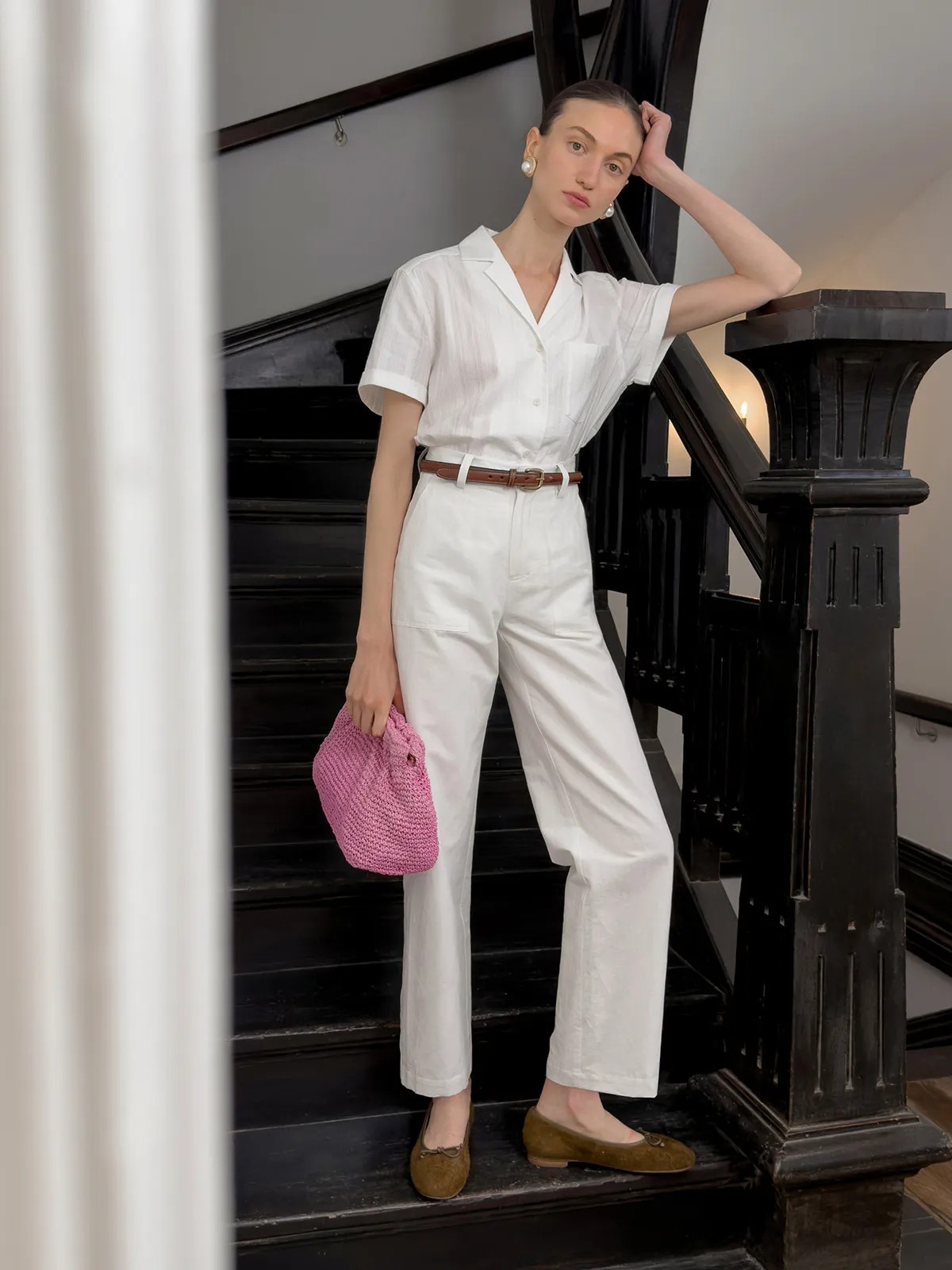 Model wearing white REID linen-cotton cropped trousers with short-sleeve shirt, brown belt, and pink crocheted bag on black staircase