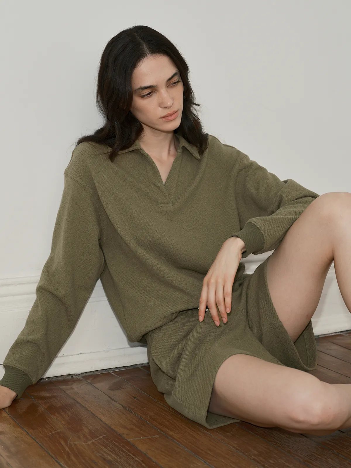 Model in olive CASPER sweatshirt and shorts seated on hardwood floor against white wall