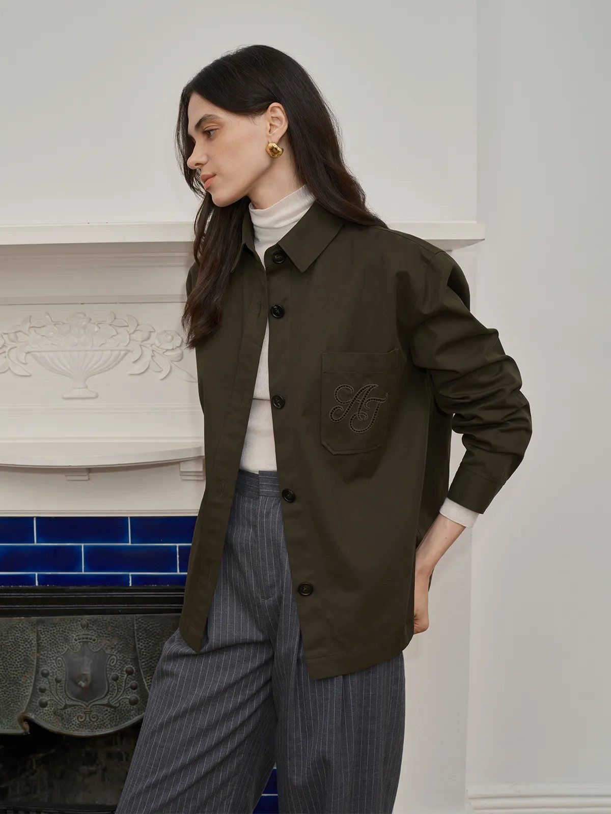 Side profile of model in HUCK jacket over white turtleneck, grey pinstripe trousers, gold earrings, by ornate fireplace