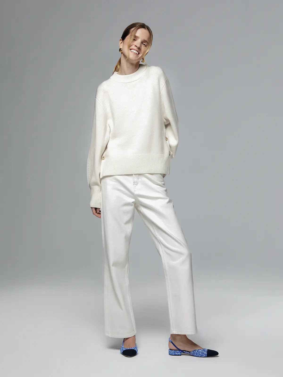 Model wearing a white merino wool sweater with metal buttons and ribbed hem, paired with white trousers.