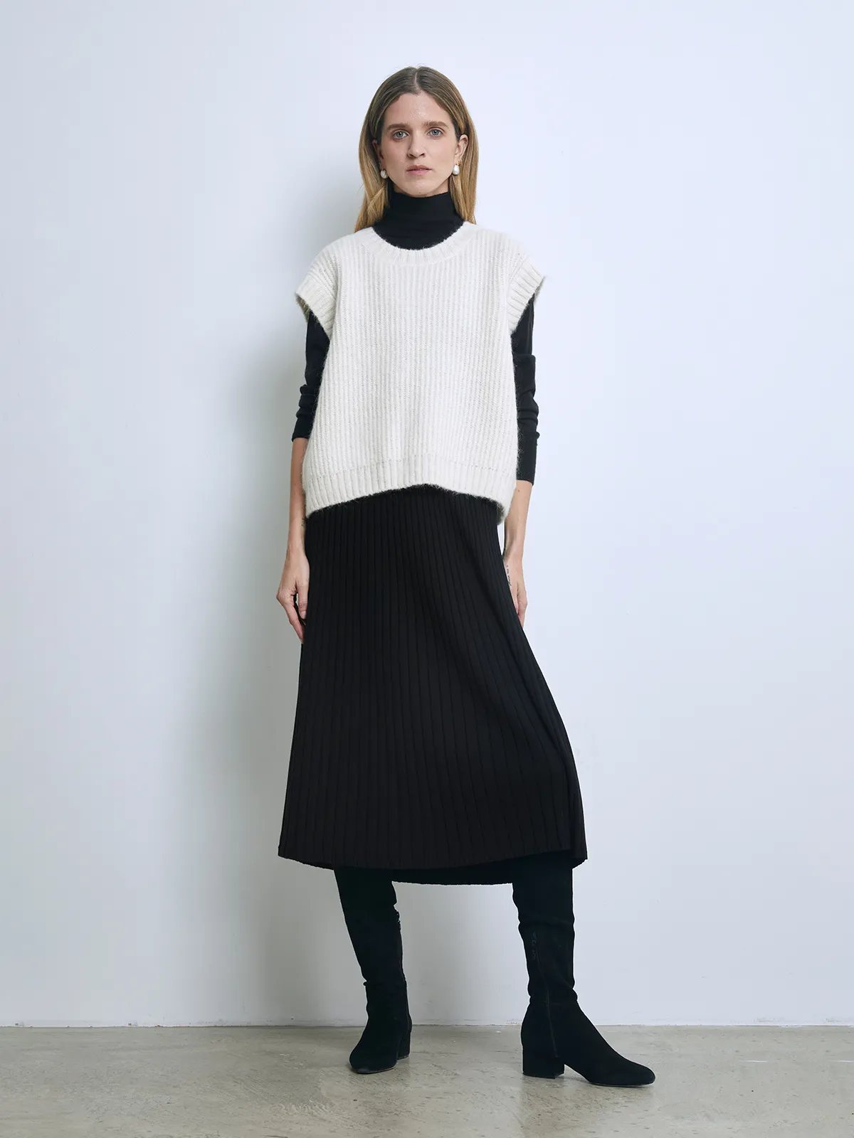 Front view of a model wearing an off-white loose boxy ribbed knit vest over a turtleneck and pleated skirt.
