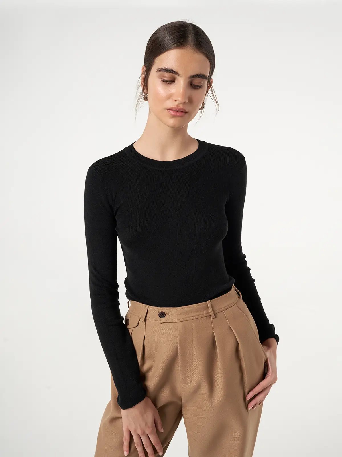 Model wearing a slim-fit, long-sleeve knit top in Black with hands resting on pleated trousers.