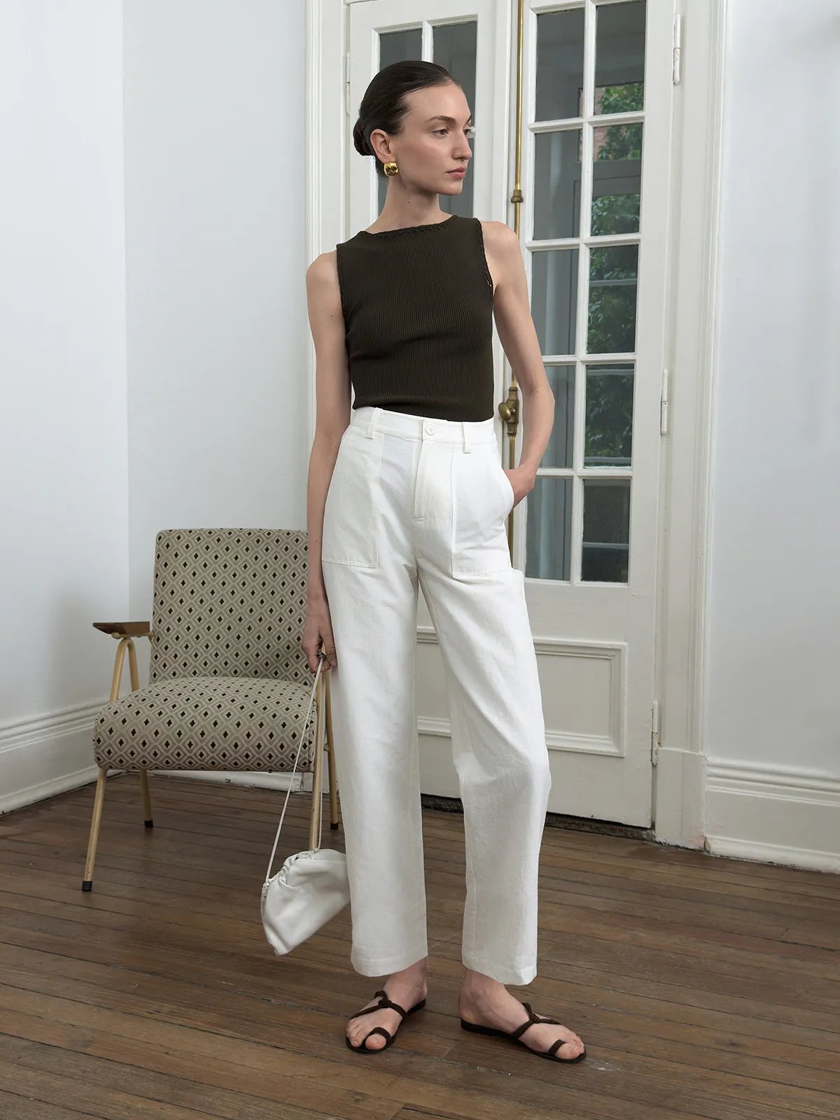 Side profile of model in white REID cropped trousers, olive sleeveless top, and white satin pouch bag near French door