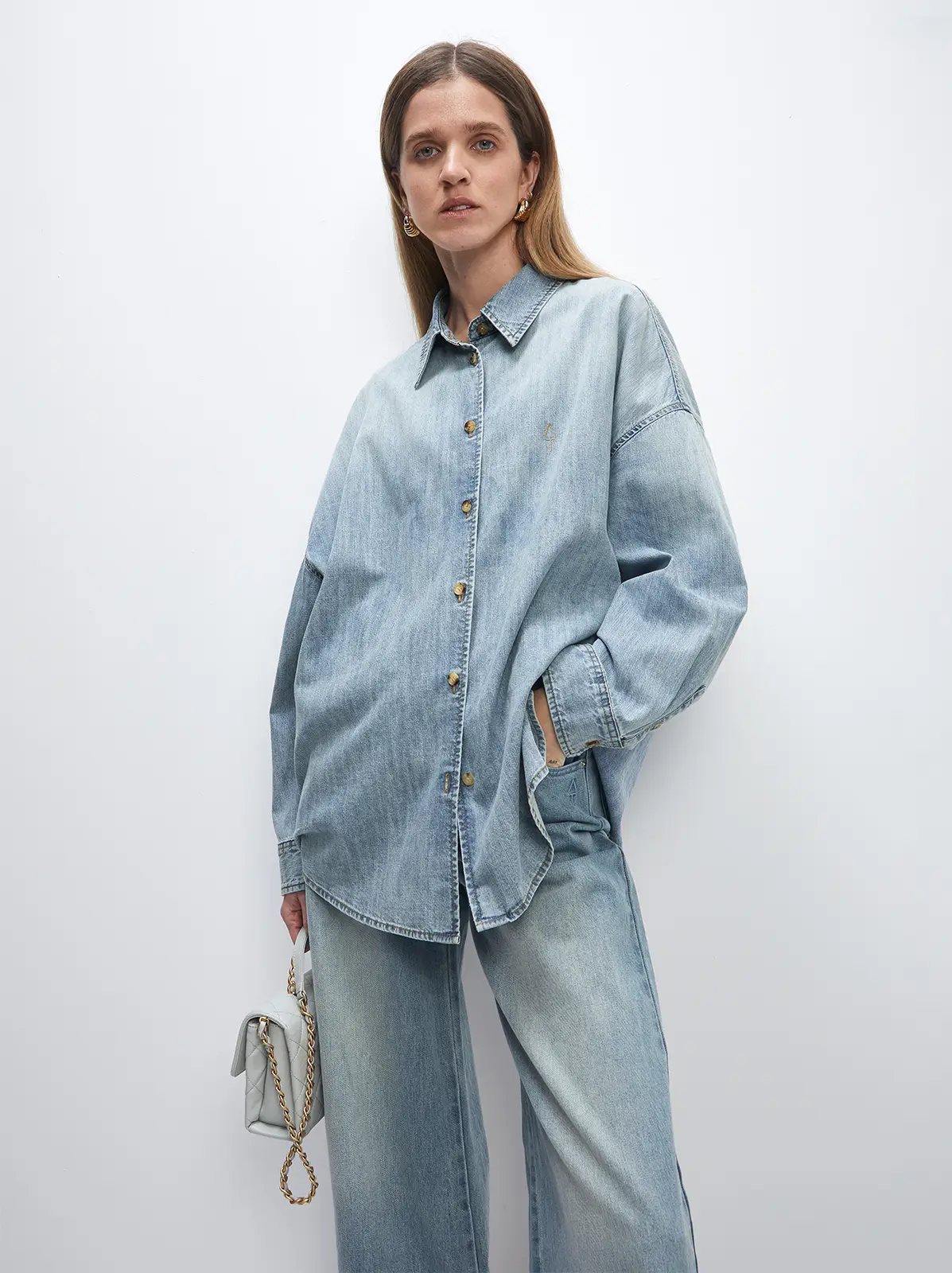 Front view of a model in a light blue oversized denim shirt with button closure, one hand in pocket, wearing matching jeans.