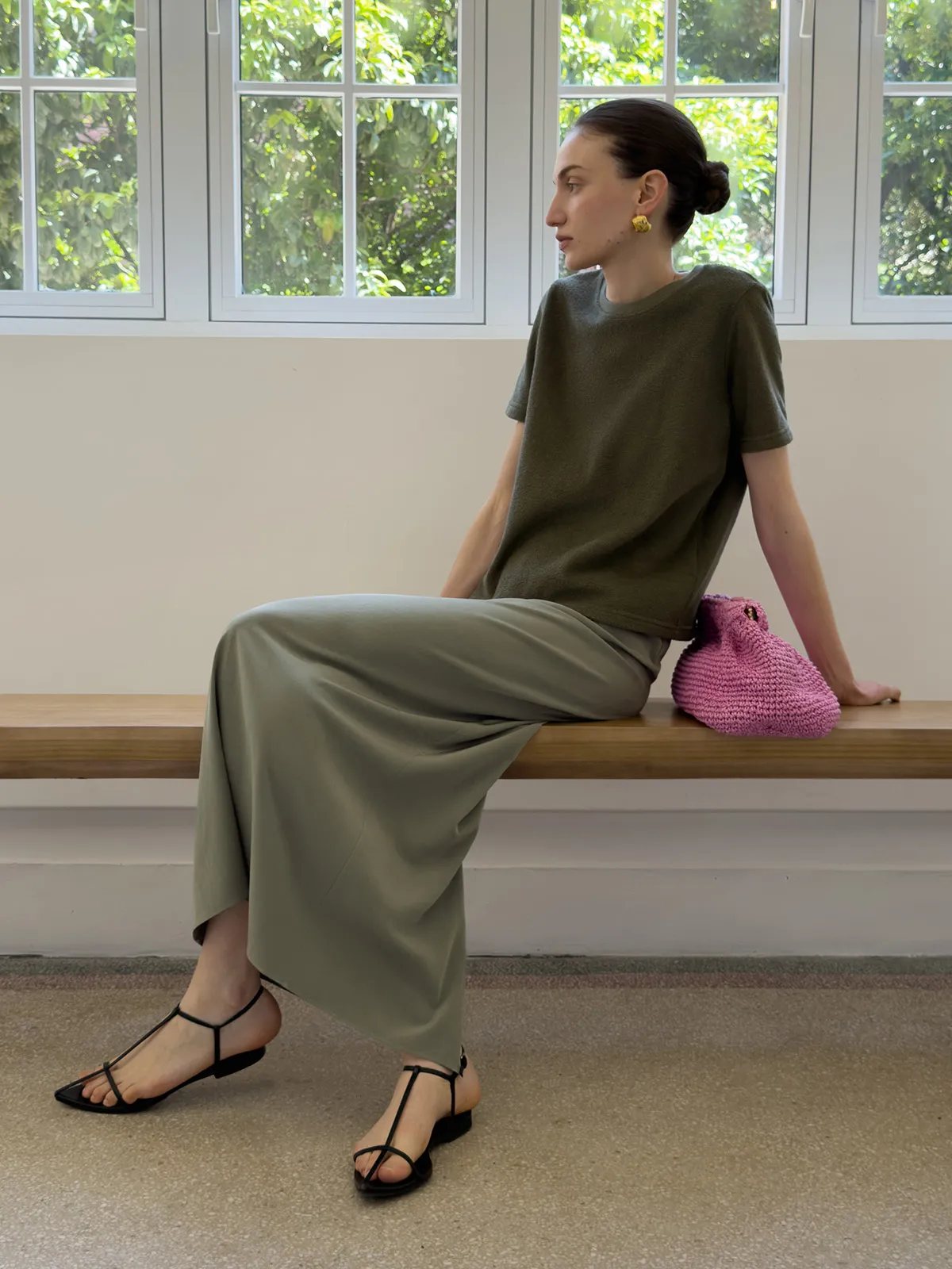 Model wearing KATRINA midi skirt with olive tee and black strappy sandals on wooden bench by window