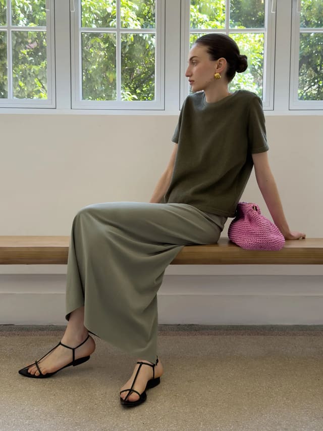 Model wearing KATRINA midi skirt with olive tee and black strappy sandals on wooden bench by window