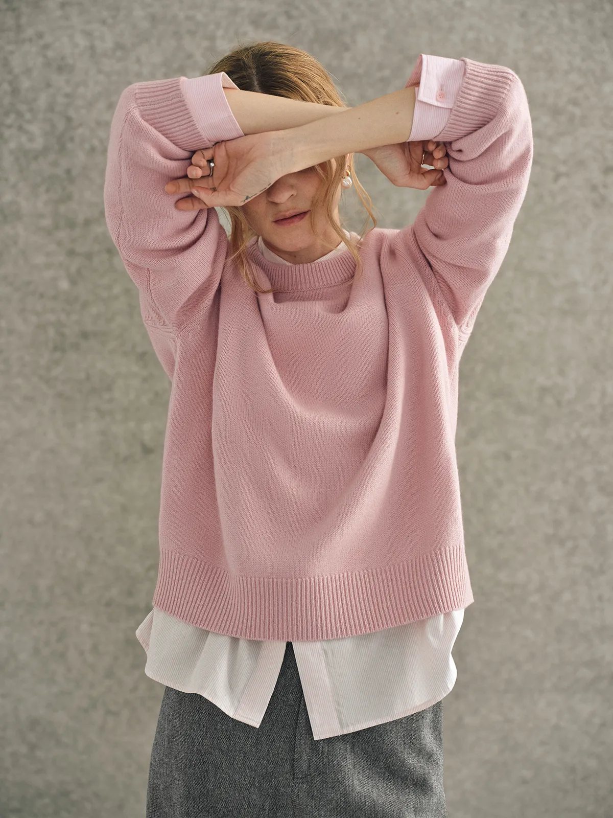 Model playfully covering eyes with arms raised, wearing pink JILL jumper over white shirt and grey trousers