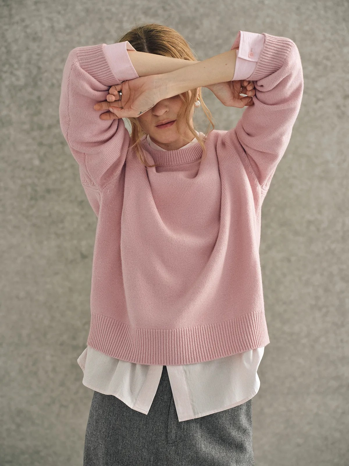 Model playfully covering eyes with arms raised, wearing pink JILL jumper over white shirt and grey trousers