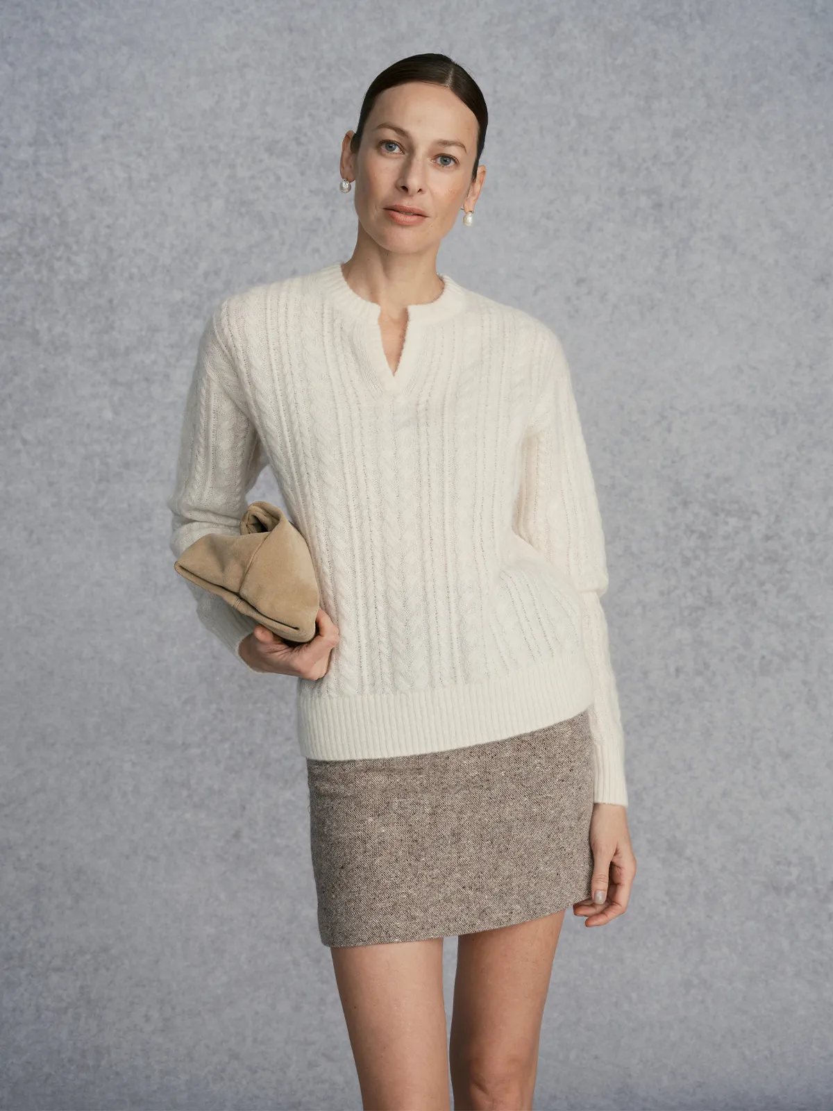 Model in KATIE skirt with ivory cable-knit sweater, holding beige suede clutch, gray textured backdrop