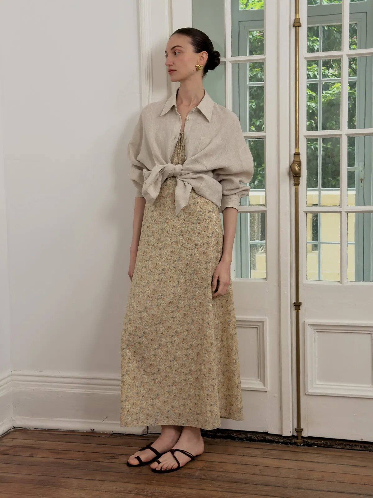 Model wearing beige floral PIPPA dress layered under oversized beige linen shirt tied at waist, near French doors