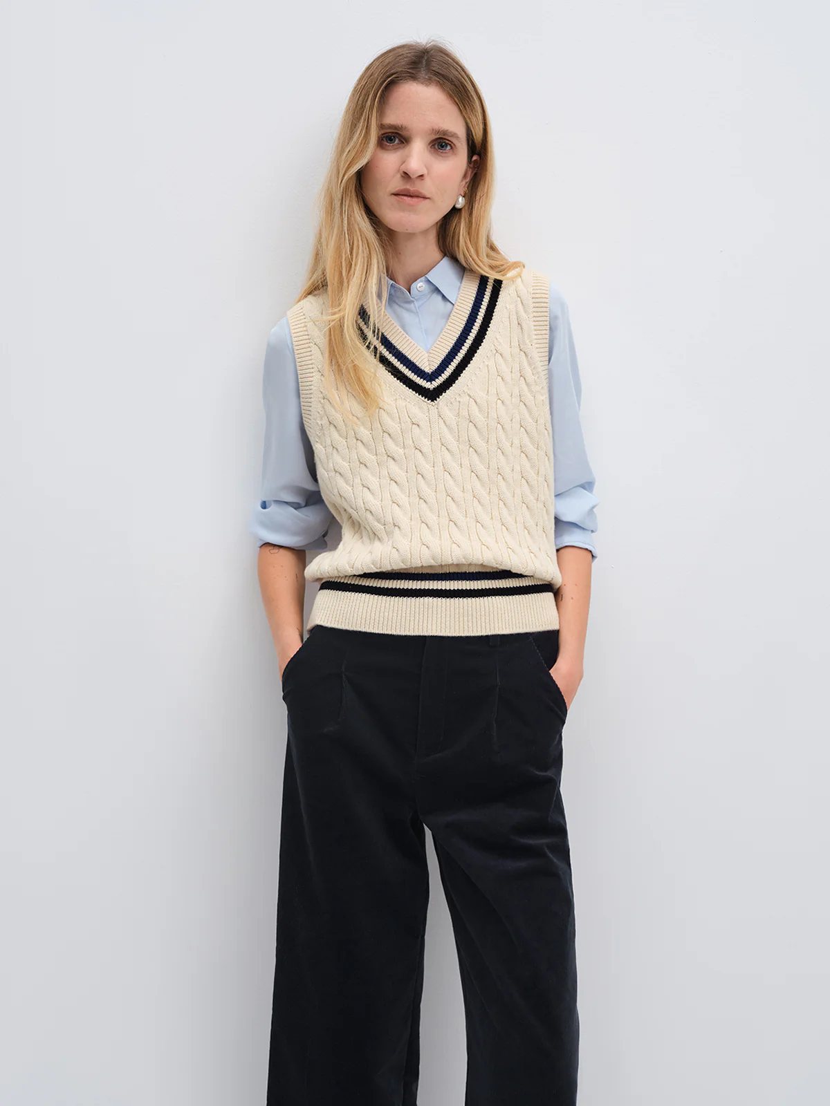 Front view of model in andreatang cream cable-knit V-neck vest with blue shirt and corduroy trousers