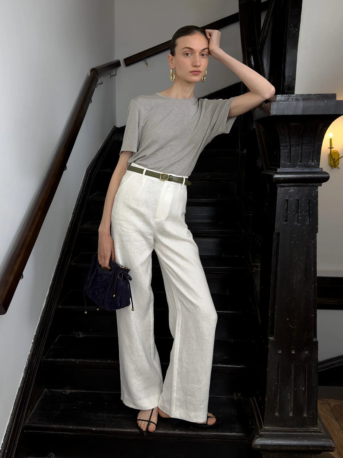 Model wearing CLORIS ivory linen trousers with grey tee, olive green belt, and navy velvet pouch on black staircase — architectural styling