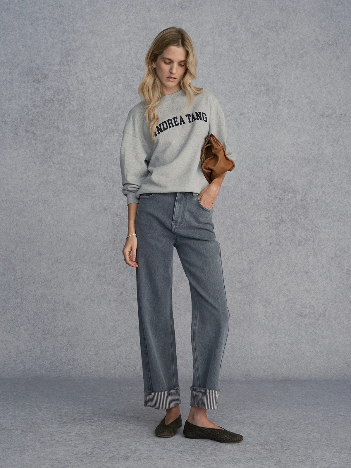 Front full-length view of model in FLIPSIDE jeans, grey sweatshirt, holding tan leather clutch