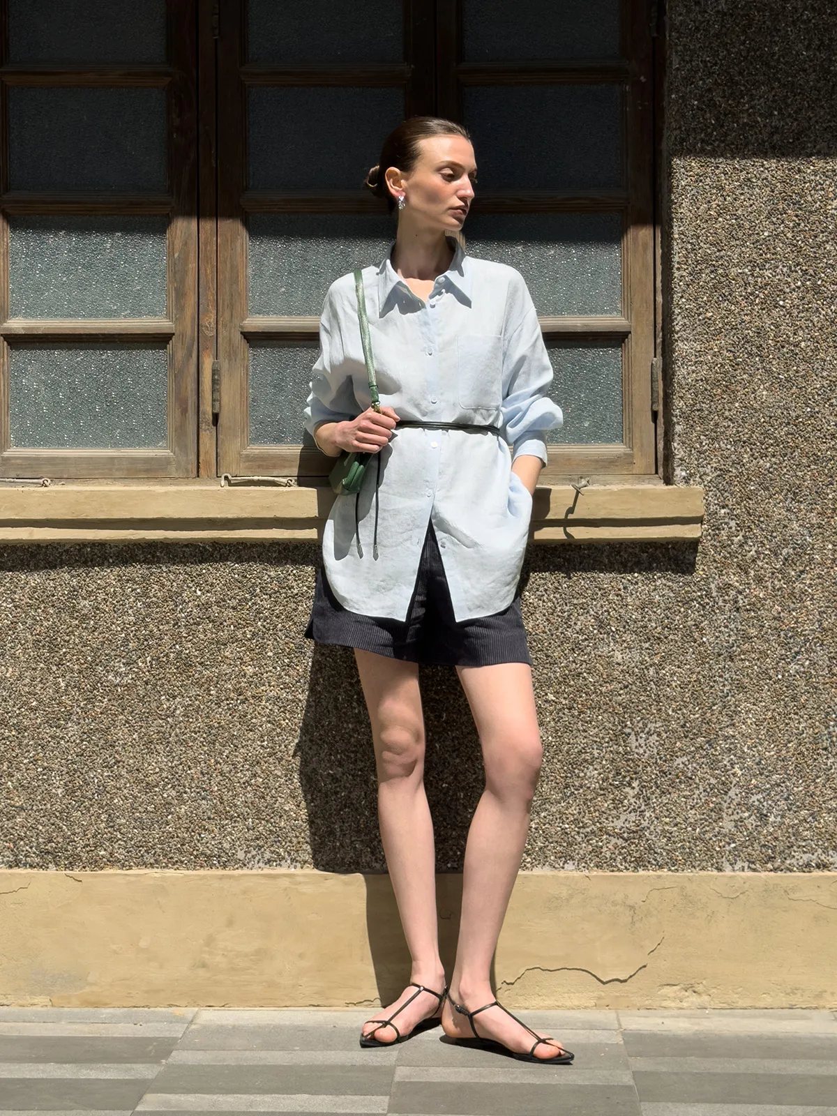 Model in light blue linen shirt belted over black striped shorts, outdoors against rustic wall