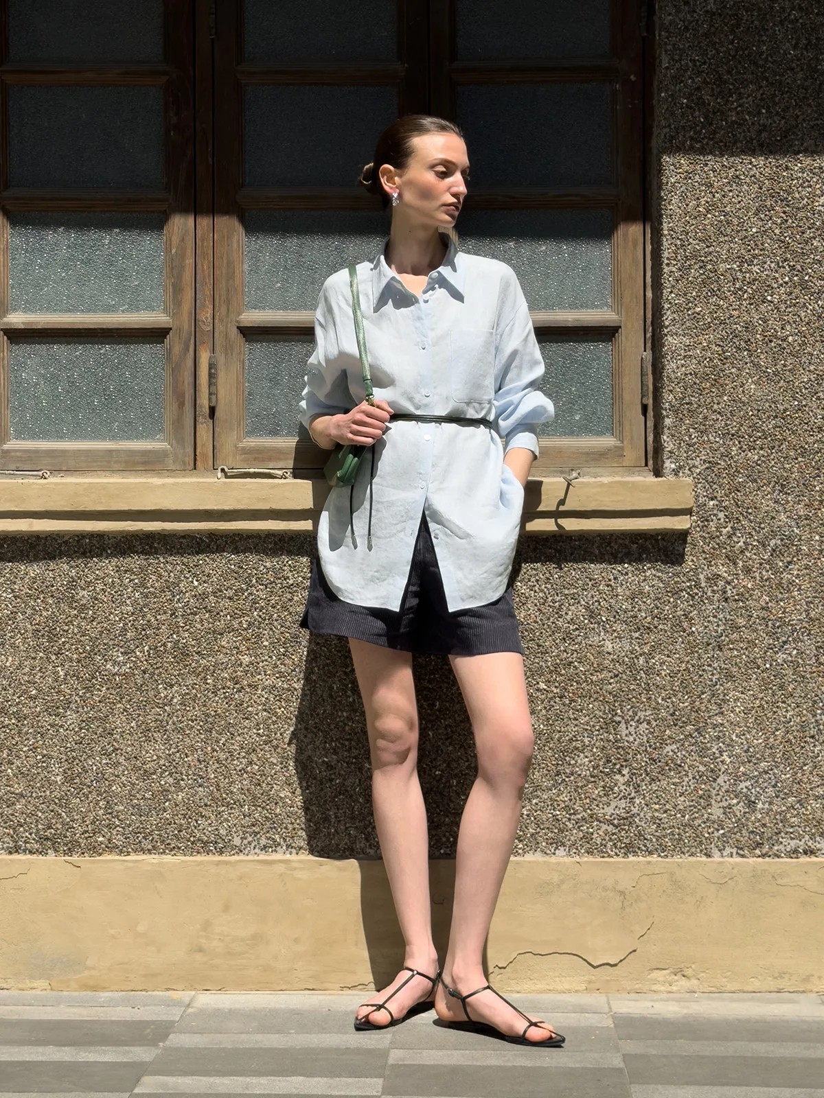 Model in light blue linen shirt belted over black striped shorts, outdoors against rustic wall