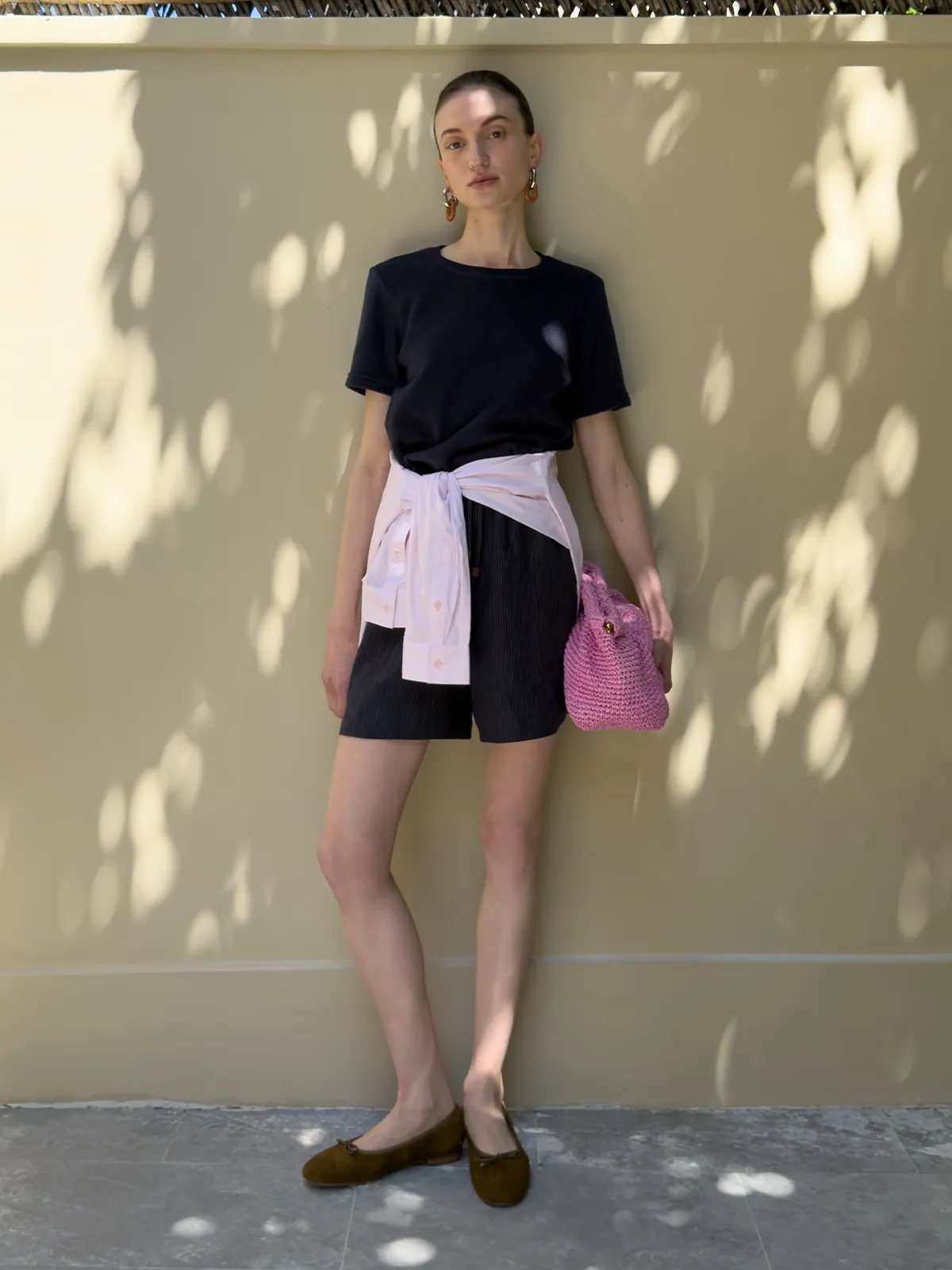 Navy LANE Terry tee styled with black pleated shorts, tied white shirt, and pink crocheted bag against sun-dappled wall