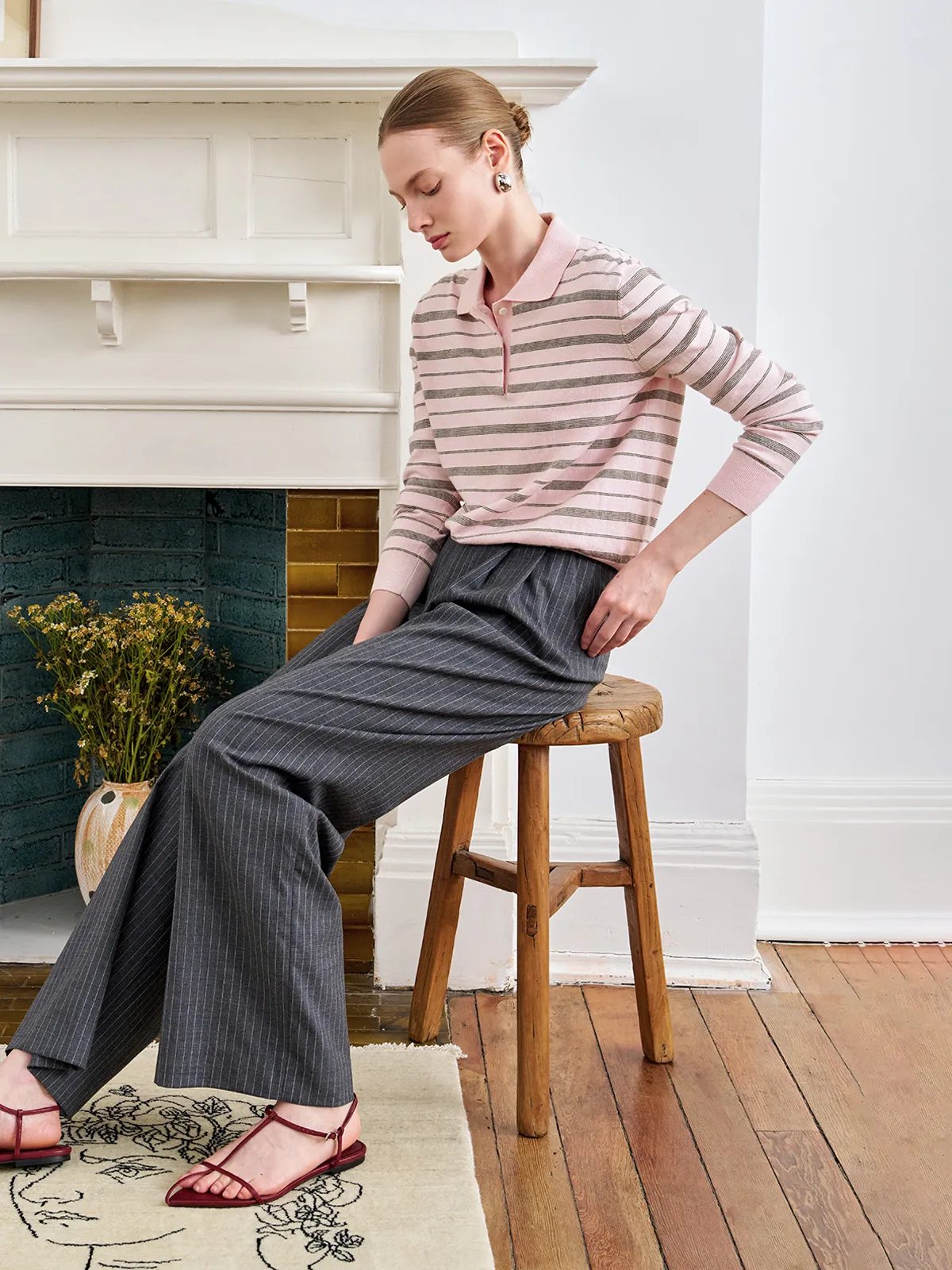 Model seated on wooden stool wearing HELENA striped polo top with wide-leg pinstripe trousers and red sandals