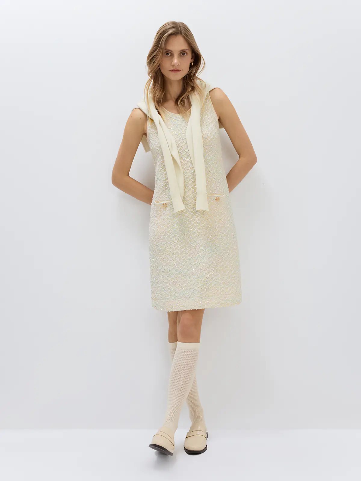 Front view of a model wearing the Beige sequin-embellished sleeveless dress with two front patch pockets and a dark heart-shaped bag.