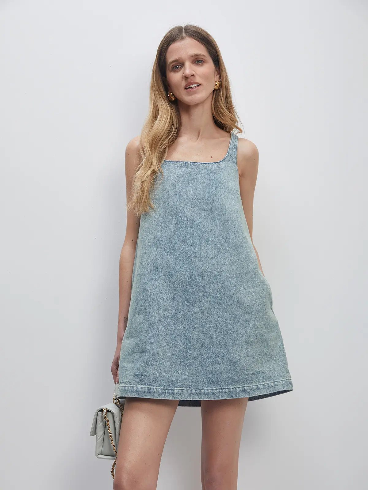 Front view of a Sky Blue retro washed A-line denim dress worn alone with a small chain-strap bag at side.